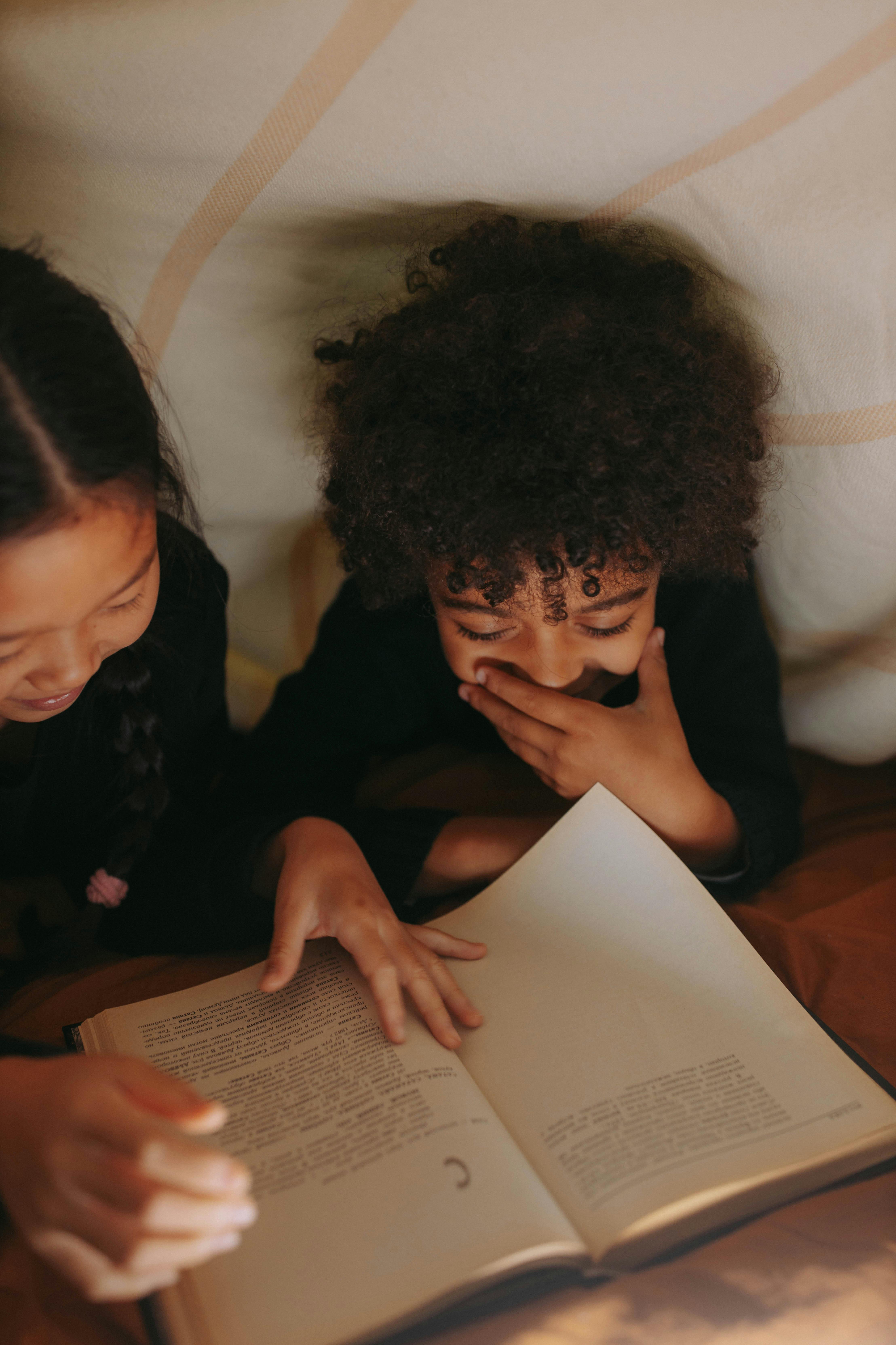 Two children reading together under a cozy blanket, laughing and connected.