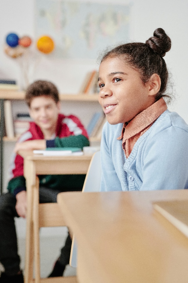 A confident Black girl smiling and engaged at her school desk, with a classmate visible in the background.