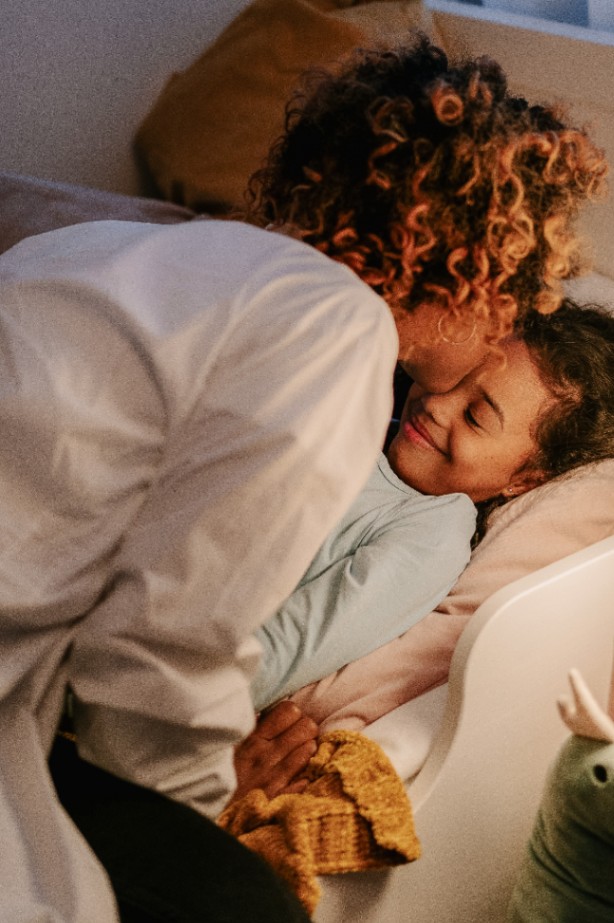 A mother with curly hair leaning over her daughter at bedtime, the child smiling with eyes closed in warm evening light.